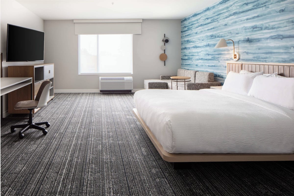 A hotel room with a large bed, a desk with a TV, a chair, a small table, and a wall-mounted lamp.