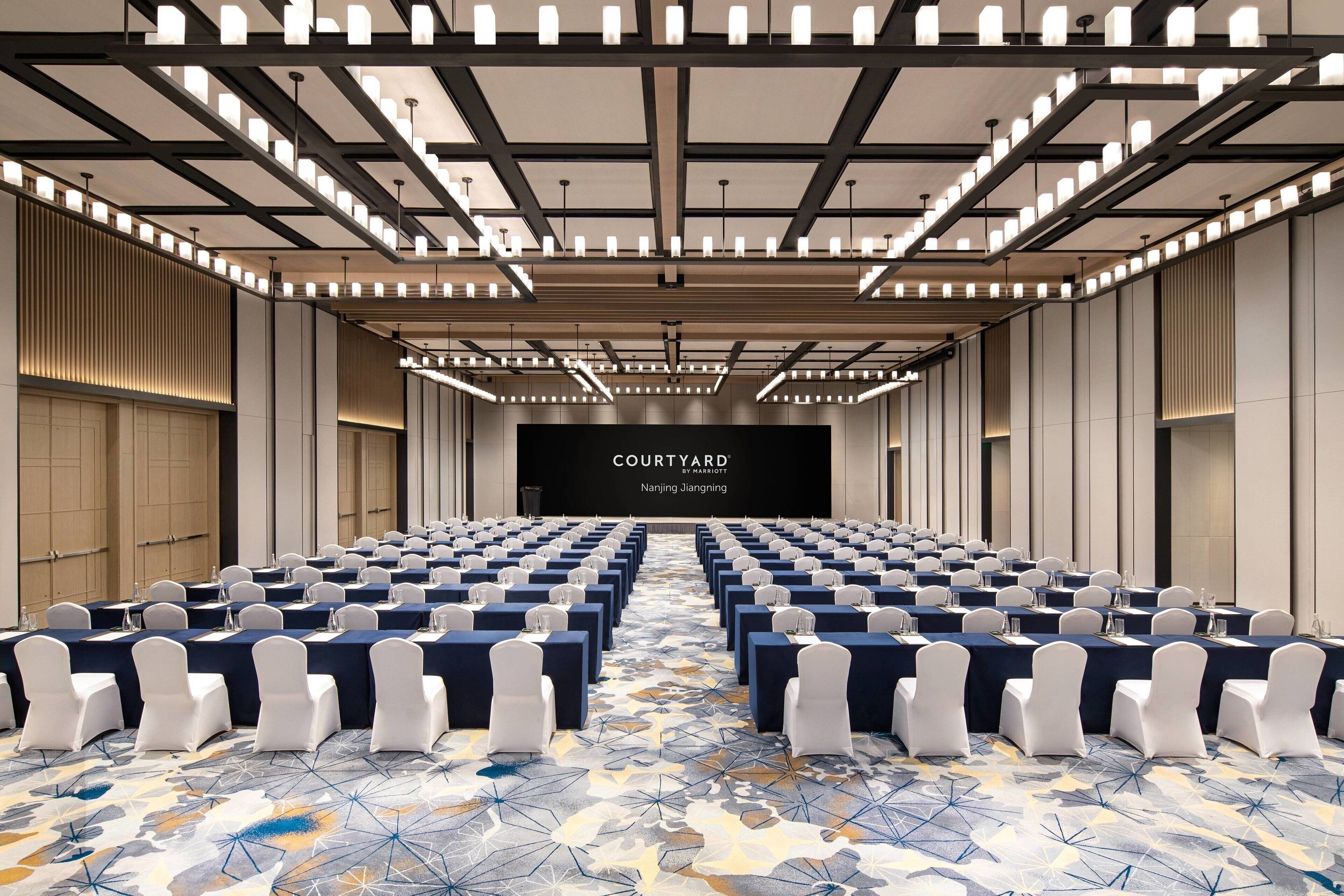 Un salón de conferencias con filas de mesas y sillas, una pantalla grande al frente y una alfombra estampada.