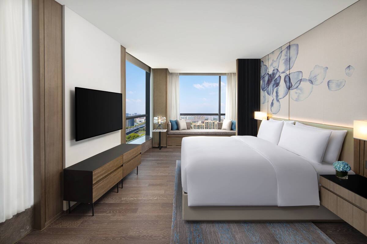 Ein modernes Hotelzimmer mit einem großen Bett, einem flachbildfernseher an der Wand, einer Bank und Blick auf die Stadt durch große Fenster.
