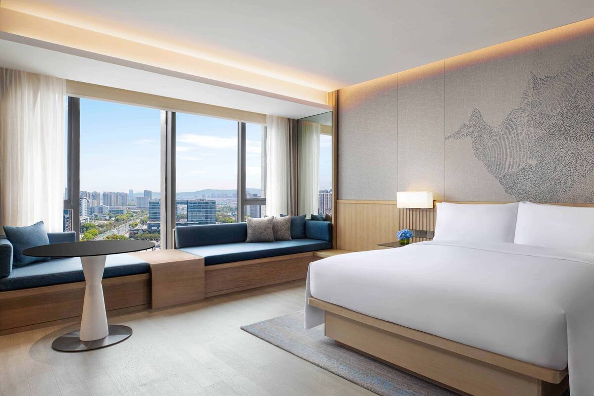 Ein modernes Hotelzimmer mit einem großen Bett, einer Sitzecke mit Blick auf die Stadt und einem großen Fenster.