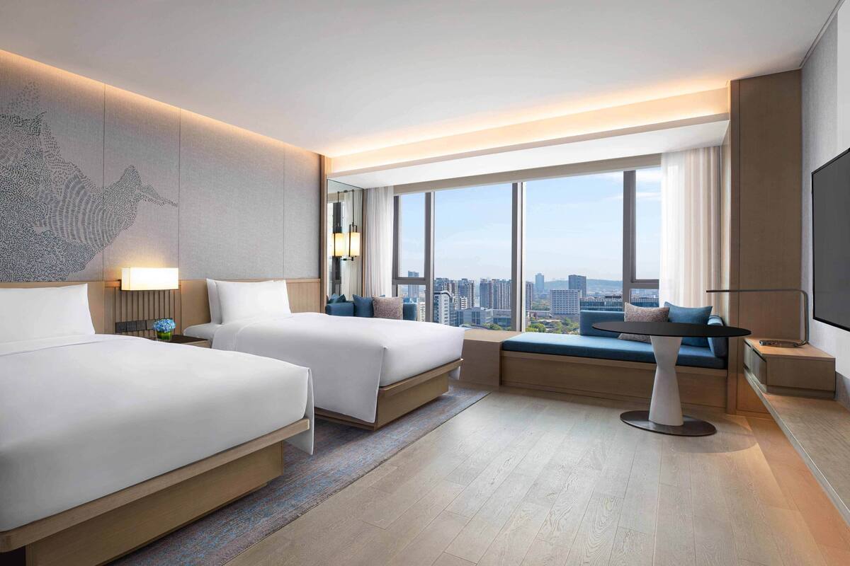 Ein modernes Hotelzimmer mit zwei Betten, einem großen Fenster mit Blick auf die Stadt, einem Flachbildfernseher und einer kleinen Sitzecke.