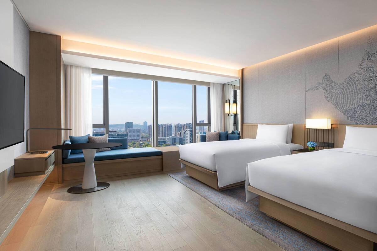 Una habitación de hotel moderna con dos camas, un sofá, una mesa de comedor y un ventanal con vistas a la ciudad.