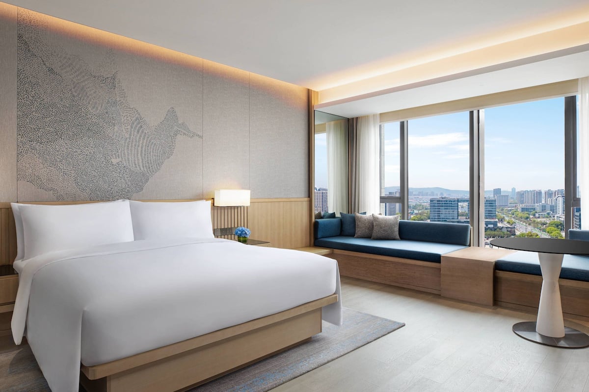 Una habitación de hotel moderna con una cama grande, un área de descanso con cojines, vista a la ciudad y una obra de arte en la pared.