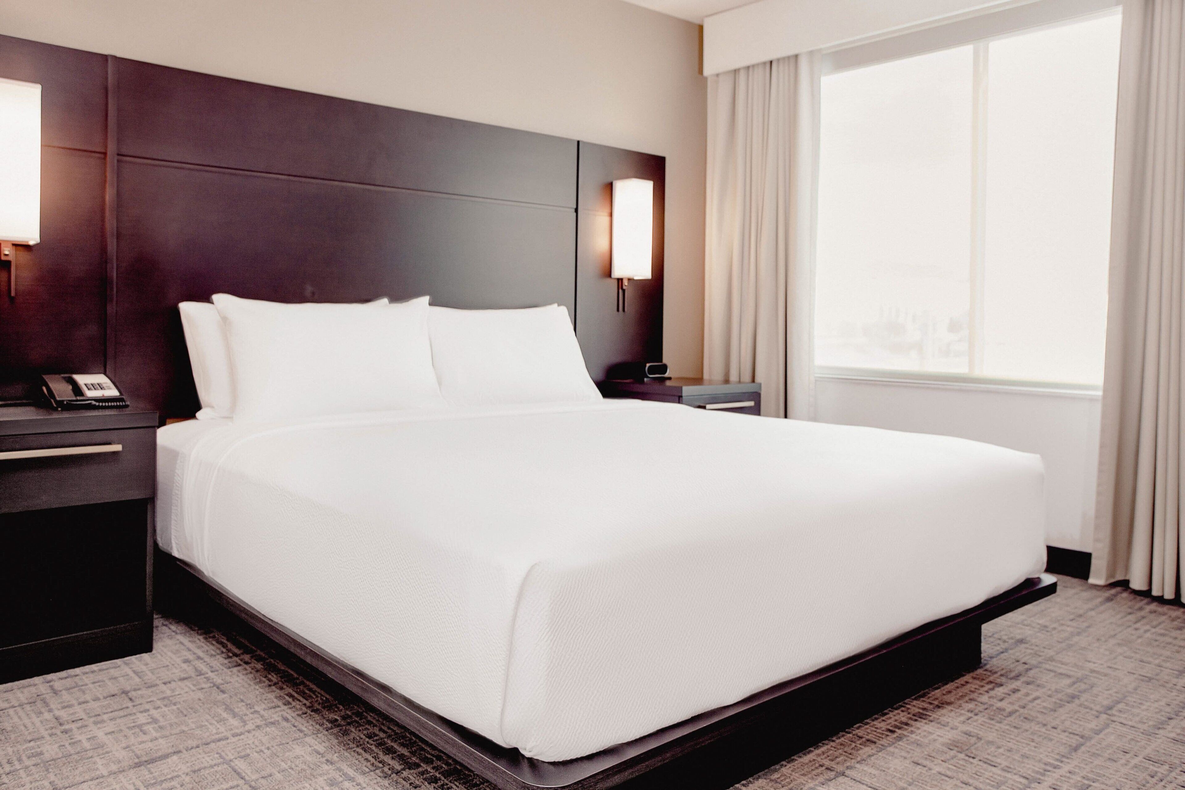 A hotel room with a large bed, dark headboard, bedside tables with lamps, and a window with curtains.