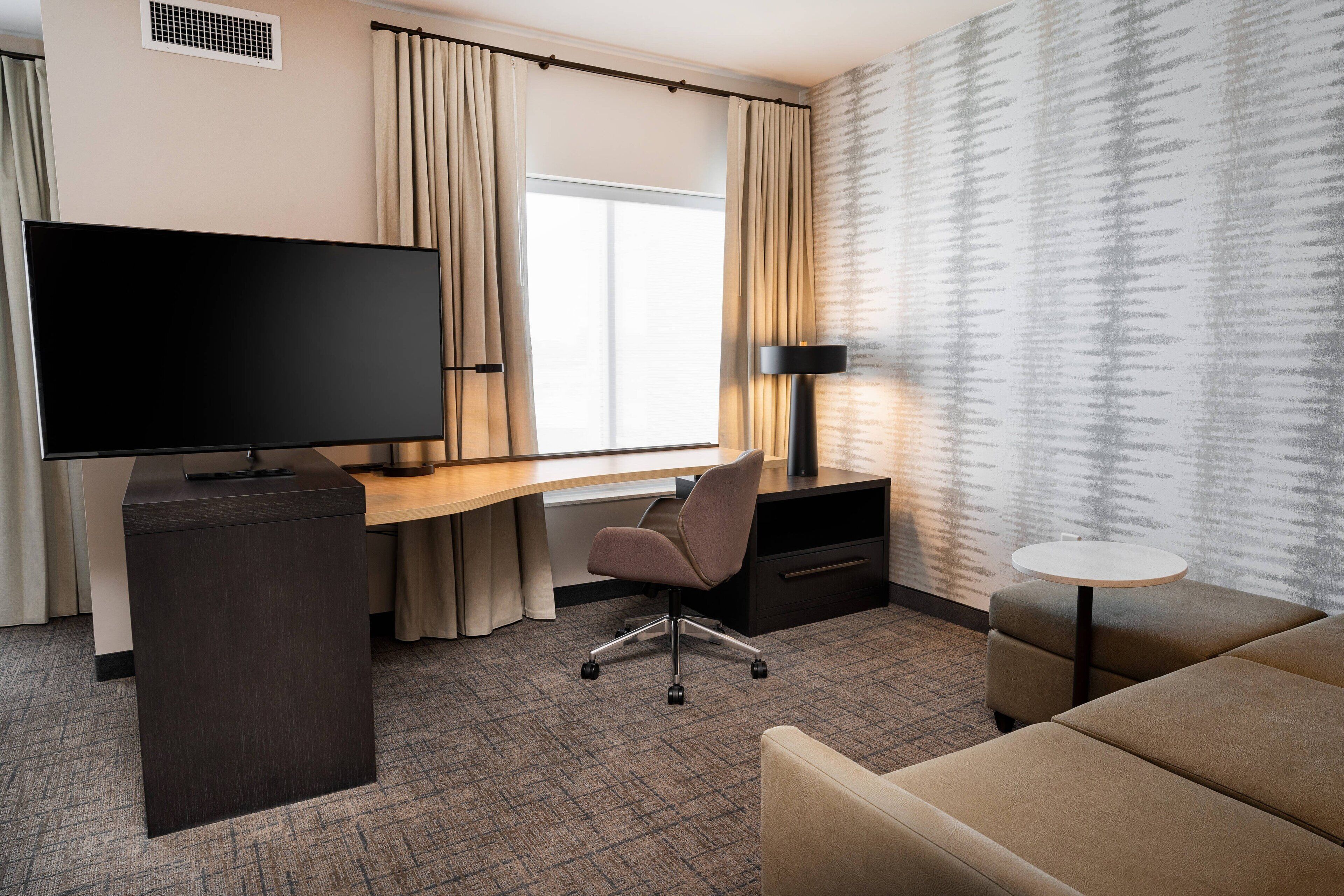 A hotel room with a flat-screen TV, a desk with a chair, a sofa, a small table, and a patterned wall.