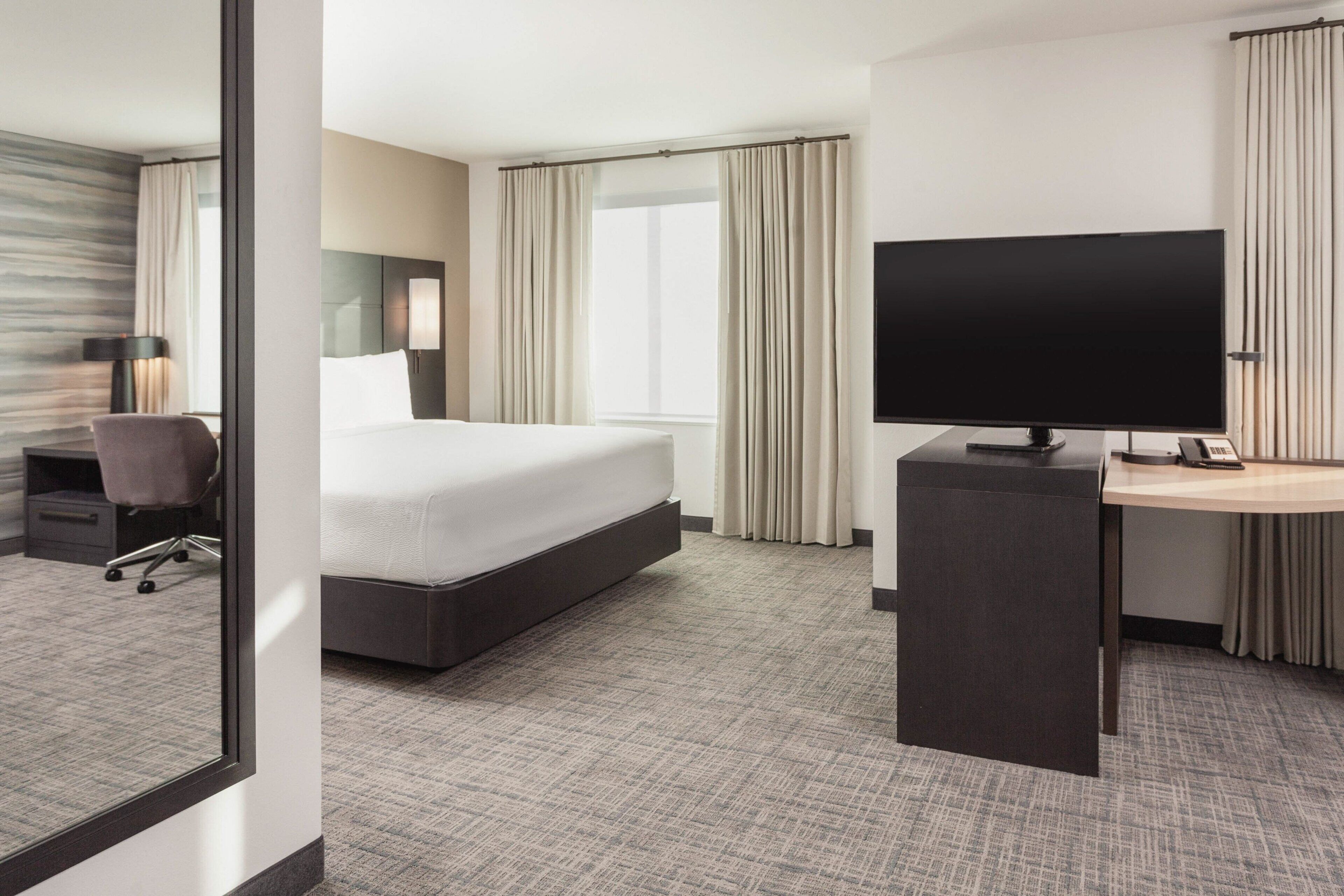 Une chambre d’hôtel comprenant un lit, un bureau avec un téléviseur à écran plat, une chaise, une lampe et une fenêtre avec des rideaux.
