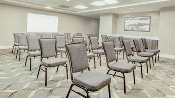 Un salón de conferencias con hileras de sillas tapizadas en gris, una gran pantalla de proyección blanca y una alfombra estampada.