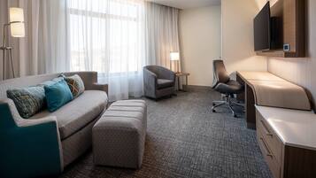 A hotel room with a sofa, two armchairs, a desk, a flat-screen TV, and a large window with sheer curtains.