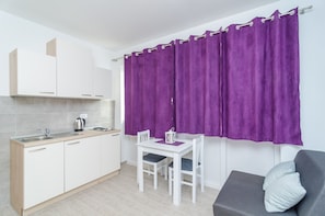 A modern kitchen with white cabinets, a sink, and a kettle. There is a dining table with chairs and a purple curtain covering the window. A gray sofa with a pillow is in the corner. - Apartments Marijan - One Bedroom Apartment (Ground Floor) - Apt 7 (Dubrovnik)