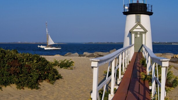 Un phare auquel on accède par une passerelle en bois, un voilier à l’arrière-plan et un ciel bleu dégagé.