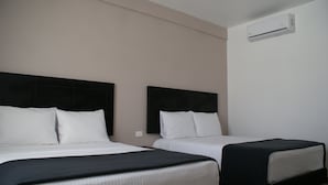 Habitación de hotel con dos camas, cada una con cabecero negro y ropa de cama blanca. Se observa una unidad de aire acondicionado de pared sobre las camas.