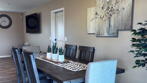 Un coin repas avec une table en bois foncé, six chaises, un ventilateur de plafond, une horloge et un tableau accroché au mur.