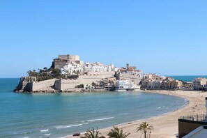 Un pueblo costero con una fortaleza destacada en una colina, una playa con gente y cielos azules despejados.