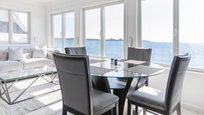 A modern dining area with a glass table and chairs, large windows with a view of the sea, and a white sofa with cushions. - PRIVATE BEACH: BEACHFRONTS & BALCONIES @ NIANTIC W/ 6 QUEENS (East Lyme)