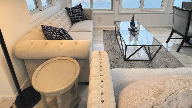 A modern living room with a sofa, ottoman, coffee table, and a lamp. There are large windows with a view of the sea. - PRIVATE BEACH: BEACHFRONTS & BALCONIES @ NIANTIC W/ 6 QUEENS (East Lyme)