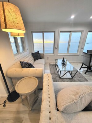 Ein modernes Wohnzimmer mit Sofa, Hocker, Couchtisch und Lampe. Große Fenster bieten Blick auf das Meer.