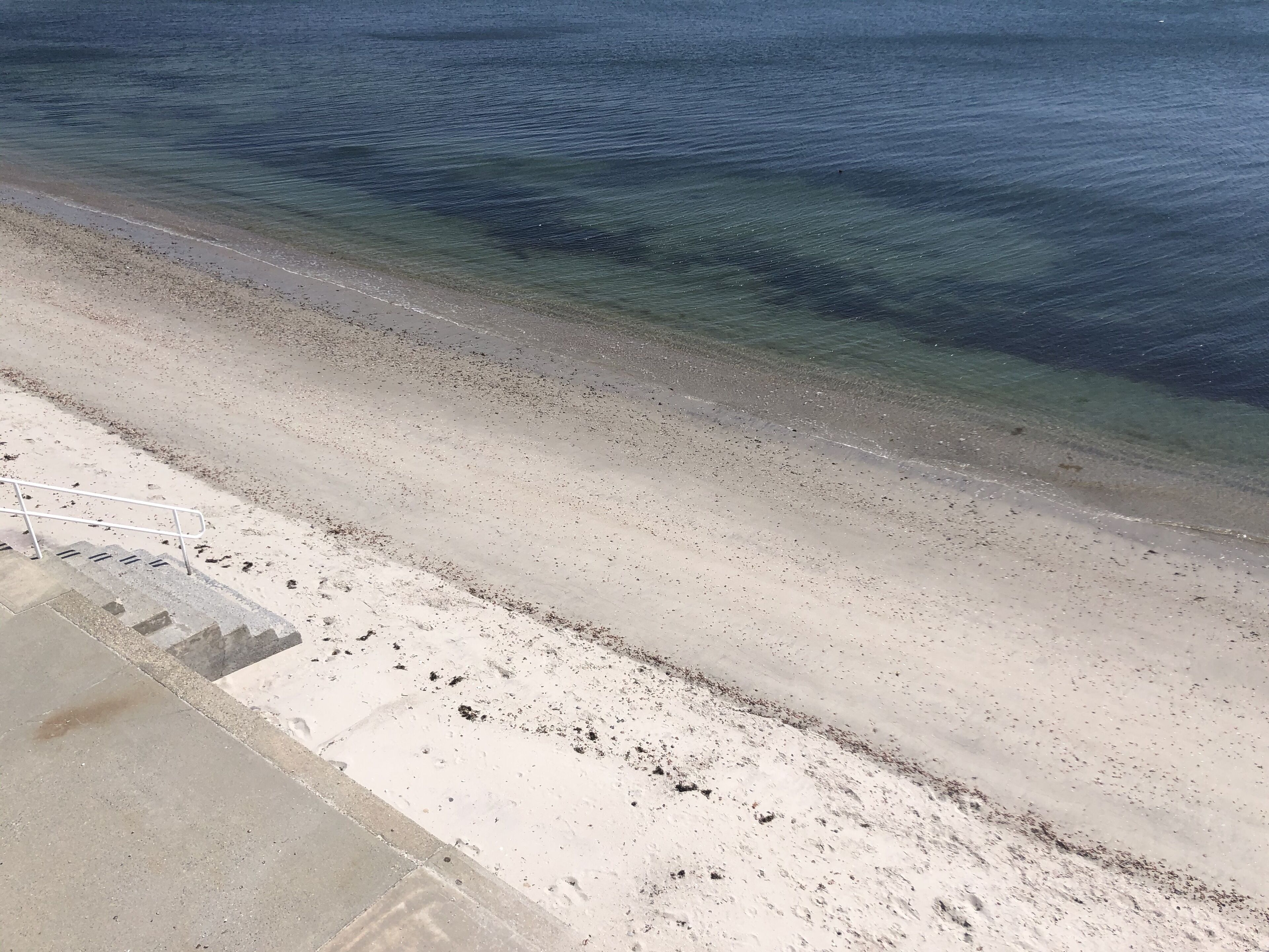 Uma praia com águas cristalinas, faixa de areia e calçada de concreto.