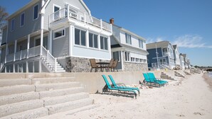 A beachfront property with a two-story house, a stone retaining wall, and a set of stairs leading to the beach. There are two blue lounge chairs on the sand. - PRIVATE BEACH: BEACHFRONTS & BALCONIES @ NIANTIC W/ 6 QUEENS (East Lyme)