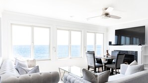 A modern living room with a large window overlooking the sea, a flat-screen TV mounted above a fireplace, and a dining area with a glass table and chairs. - PRIVATE BEACH: BEACHFRONTS & BALCONIES @ NIANTIC W/ 6 QUEENS (East Lyme)