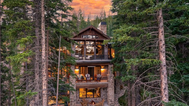 A two-story log cabin with a stone foundation, surrounded by dense forest.