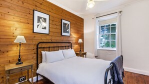 A bedroom with a wooden paneled wall, a bed with white bedding, two bedside lamps, and a window with a view of greenery.
