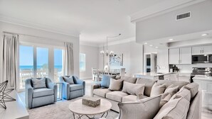 A spacious living room with a large sofa, armchairs, and a coffee table. There is a view of the ocean through large windows.