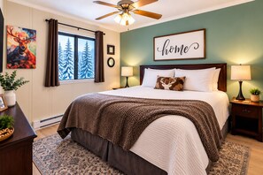 A bedroom with a large bed, a ceiling fan, a window with curtains, a wall-mounted picture, a bedside table with a lamp, and a decorative pillow. - Summit 108B | 3BR | Sunset views | Free Parking (Snowshoe)