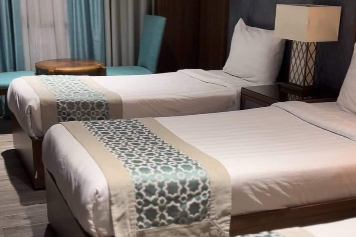 A hotel room with two beds, a wooden headboard, a nightstand with a lamp, and a chair.