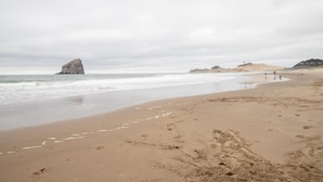 Una playa arenosa con una gran formación rocosa a lo lejos, algunas personas caminando y un cielo nublado.