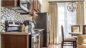 Una cocina moderna con horno empotrado, microondas y refrigerador, un comedor con mesa redonda y sillas, y un amplio ventanal con cortinas.