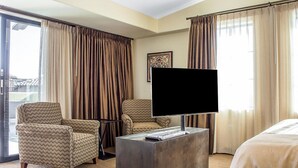 Habitación de hotel con un televisor de pantalla plana grande, dos sillones con motivos estampados, una mesa de centro con un control remoto y una cama.