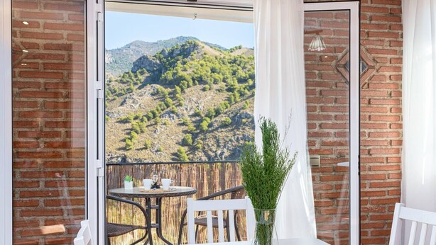 A balcony with a table and chairs, a vase of greenery, and a view of a mountainous landscape.