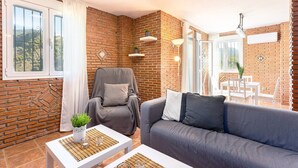 A cozy living room with a brick wall, a grey sofa, a grey armchair, and a dining area with a table and chairs.