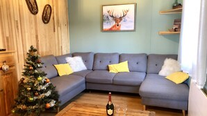 A cozy living room with a grey sectional sofa, a wooden coffee table with a bottle and glasses, a decorated Christmas tree, and a painting of a deer on the wall. - Cosy chalet close to resorts and lake many activities summer/winter 10 pl. ⛷️🥾🦌 (Matemale)