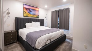 A bedroom with a bed, a nightstand, a wall-mounted lamp, a painting, and a window with curtains.