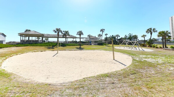 Una cancha de voleibol de arena con red, rodeada de césped y palmeras.
