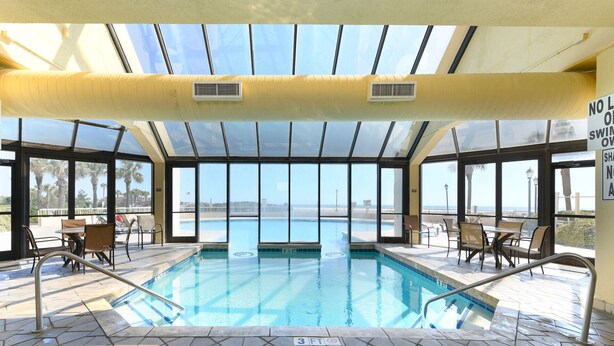 An indoor swimming pool with clear views of the ocean, surrounded by sunlit patio seating and a glass ceiling.