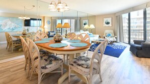 A spacious living area with a round wooden dining table surrounded by wicker chairs, a large window with a view of the city, and a modern chandelier.