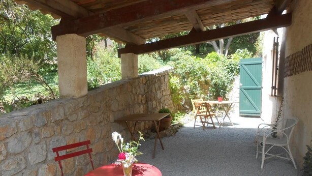 Un patio al aire libre con un muro de piedra, una mesa redonda con sillas rojas y un área techada con techo de madera.