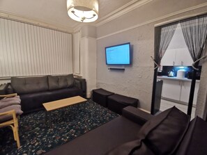 A living room with a dark leather sofa, a small wooden coffee table, a TV mounted on the wall, and a kitchen area visible through a partially open door.