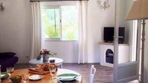 Un coin repas avec une table ronde en bois, dressée pour deux personnes, entourée de chaises blanches. Une fenêtre avec des rideaux laisse entrer la lumière naturelle dans la pièce.