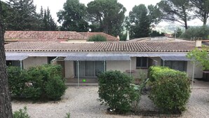 A building with a tiled roof, a covered patio area, and a gravel courtyard with various shrubs. - La Cigale Verte à Callian (CALLIAN)