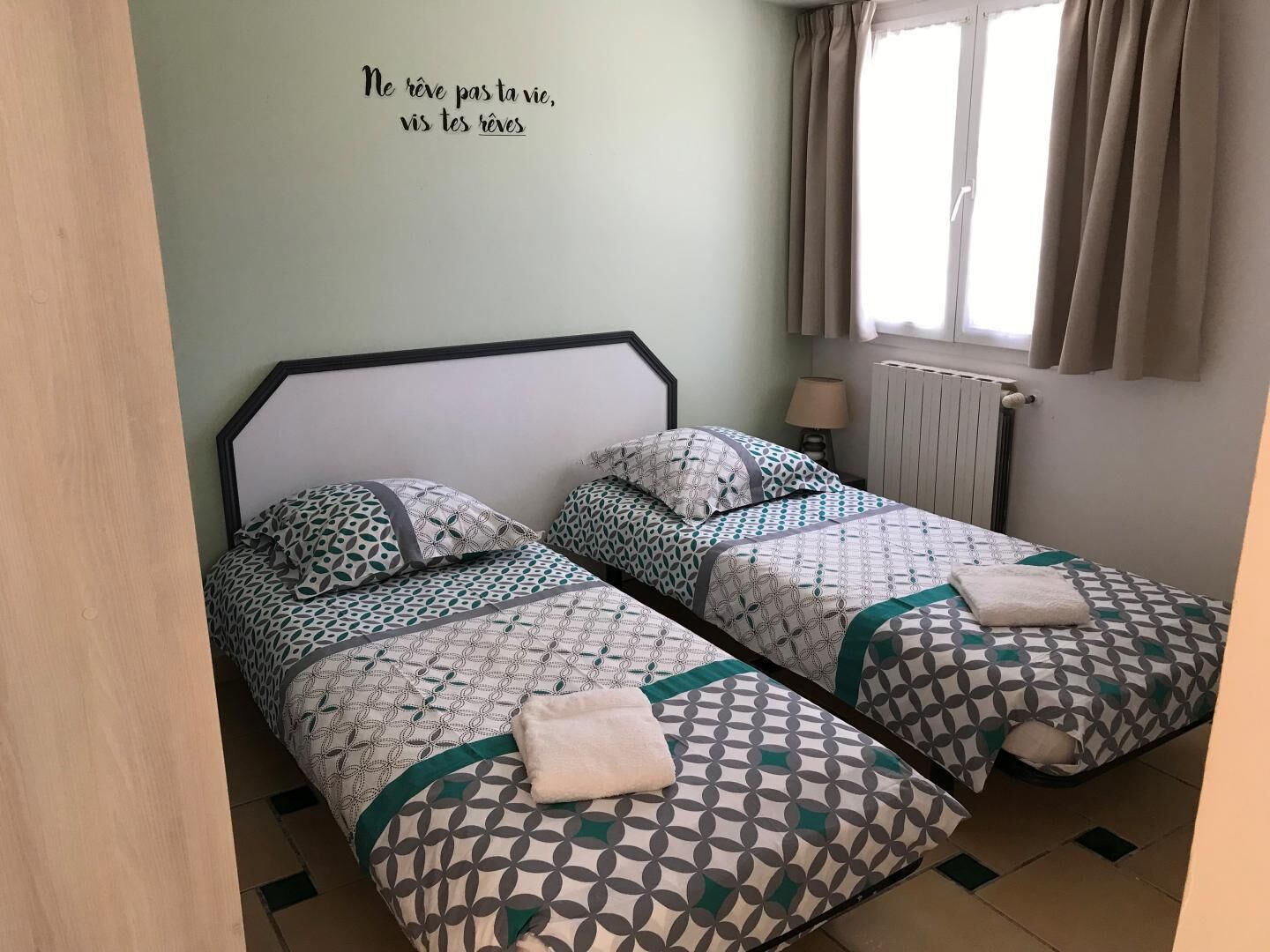 A room with two beds, each with patterned bedding and a towel on the bed. There is a wall with a quote in French, a window with curtains, and a radiator.
