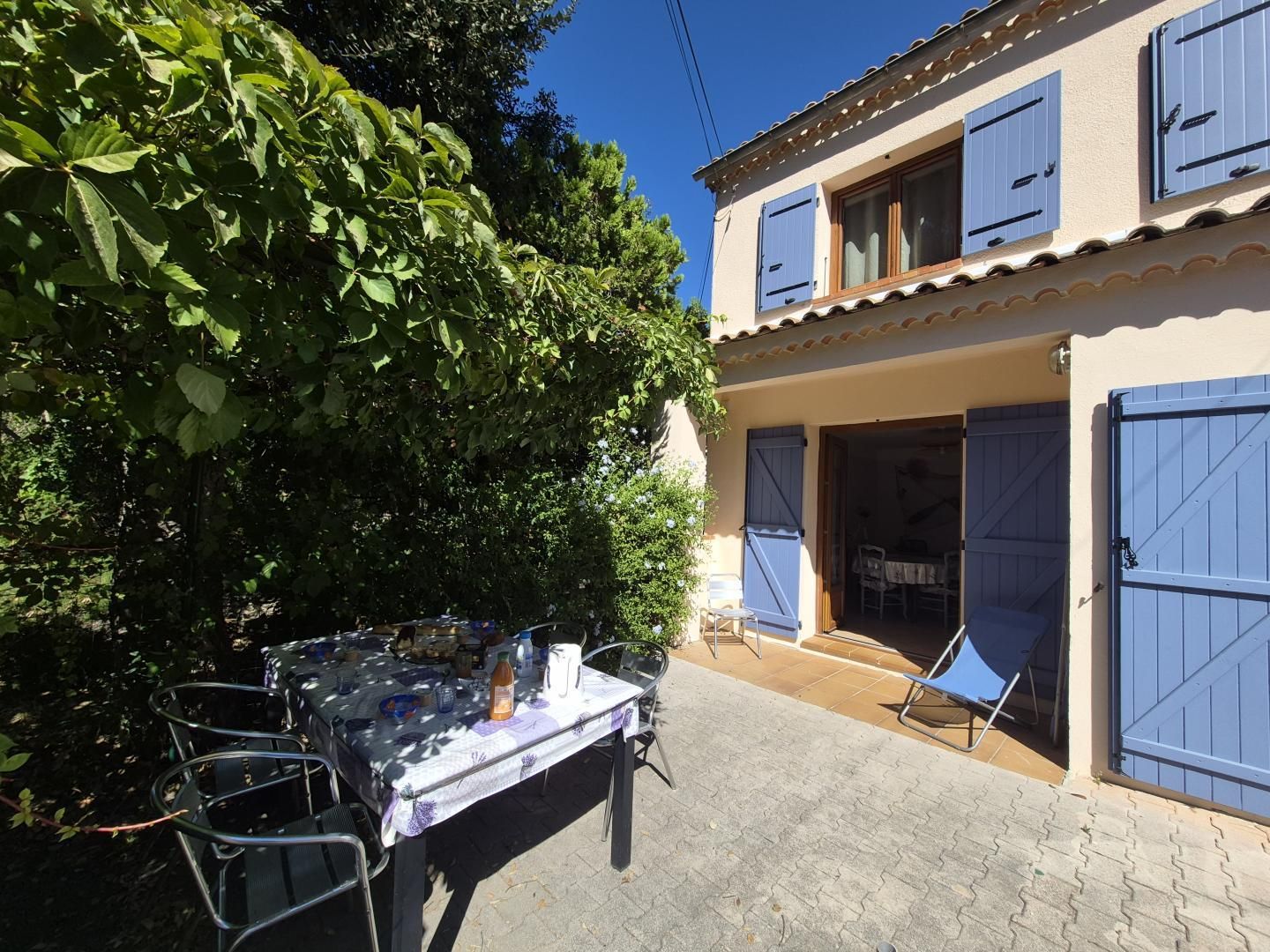 A patio with a table set for dining, surrounded by chairs, and a building with blue shutters and a tiled roof.