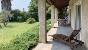 Ein überdachter Patio-Bereich mit Liegestuhl, Tisch und Stühlen und Blick auf eine gepflegte Rasenfläche und Bäume.