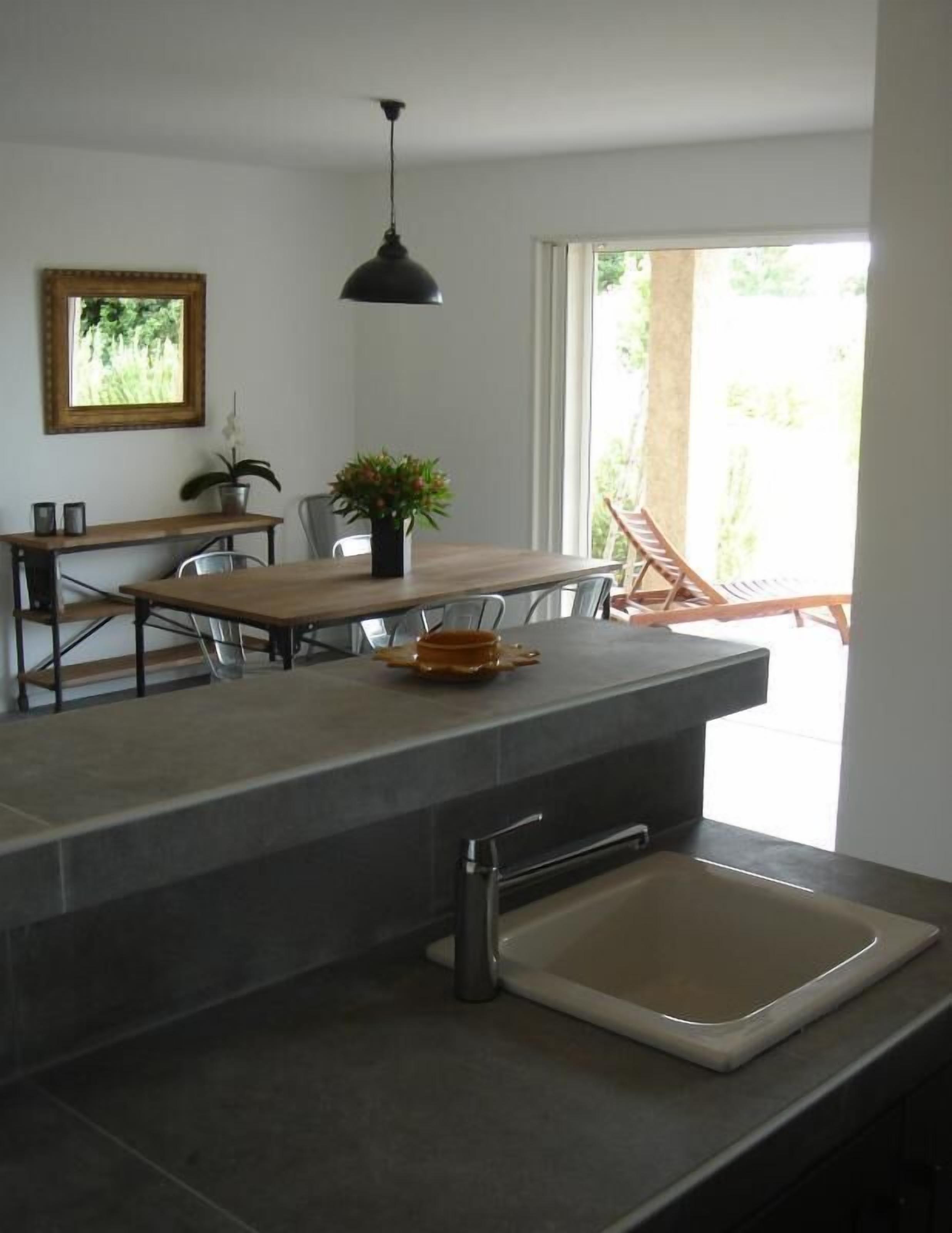 A modern kitchen with a large island, a dining area with chairs, a hanging light fixture, and a view of the outdoors.