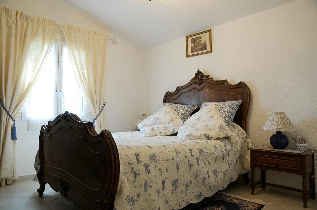 A bedroom with a wooden bed, floral bedding, a nightstand with a lamp, and a window with curtains.