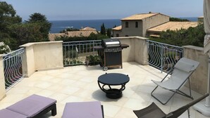 Un balcón con una parrilla, sillones de descanso y una mesa, con vistas a la costa y edificios de tejas rojas.