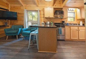 A wooden cabin interior with a kitchen, two teal armchairs, a small dining table, and a TV mounted on the wall.