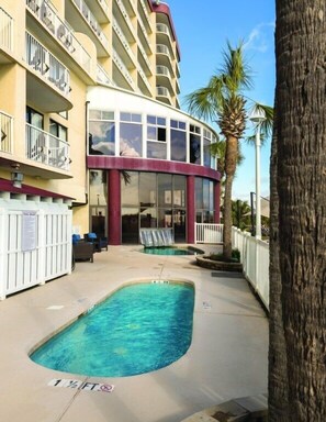 A pool area with a building in the background, a palm tree, and a sign indicating a 1 1/2 ft depth. - North Myrtle Beach | Friends Beach Weekend | Pool + Beach | 2BR (North Myrtle Beach)